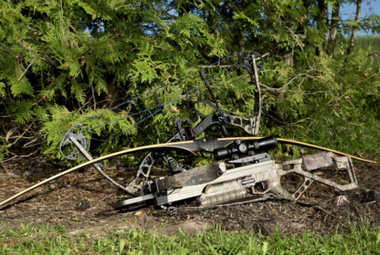 Camouflaged hunting rifle and bow lay in the dirt on the ground in front of green vegetation.