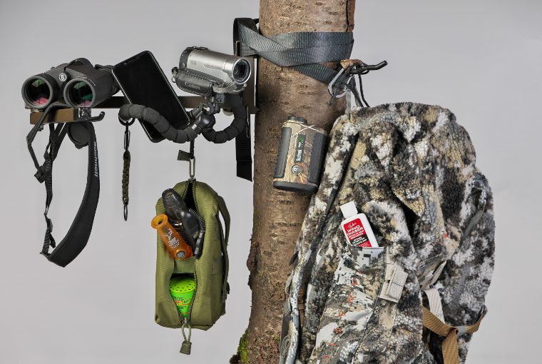 Binoculars, a black iphone, a video camera, a camouflage backpack, and other accessories hang tied on a tree.