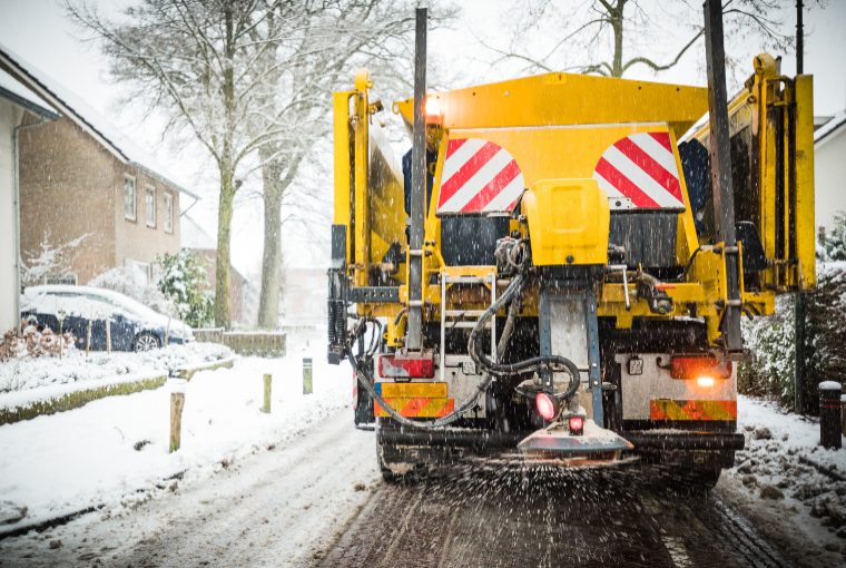 A large yellow truck disperses salt onto a residential street as snow falls on a cold-looking winter day.