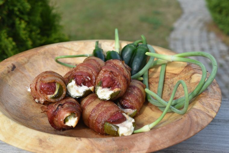 (Jalapeno dove poppers) Crispy looking bacon wrapped around cream cheese, jalapeno, and dove set on a wooden plate alongside green vegetables.