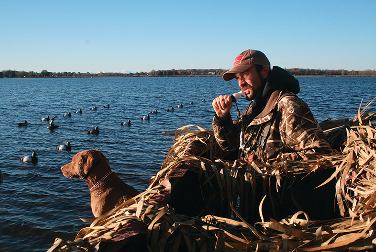 a waterfowl'er sits, calling in his boat amongst the decoys, with his pup at the ready